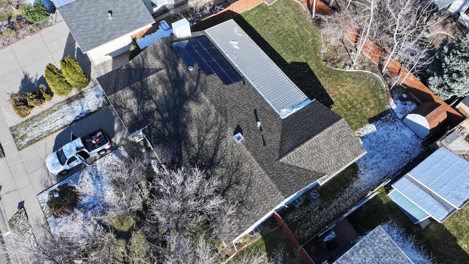 Top-down view of a dark shingle roof shaded by bare trees with a dusting of snow in the yard